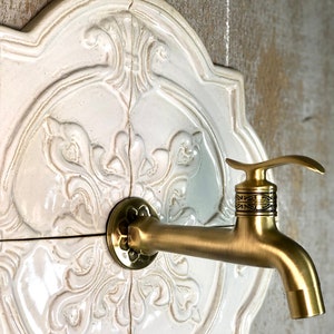 Puede incluir: Un grifo de color dorado con un diseño ornamentado está montado en una baldosa de cerámica blanca con un patrón floral decorativo.