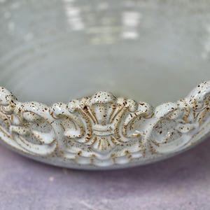 Puede incluir: Un cuenco de cer&aacute;mica blanca con un borde decorativo y ornamentado. El borde tiene un dise&ntilde;o elevado y detallado con un glaseado marr&oacute;n moteado.