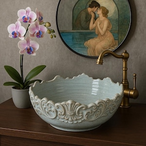Puede incluir: Un lavabo de ba&ntilde;o de cer&aacute;mica azul claro con un dise&ntilde;o ornamentado y decorativo alrededor del borde. El lavabo est&aacute; sobre una encimera de madera con un grifo dorado.
