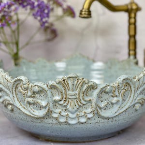 Puede incluir: Un lavabo de cer&aacute;mica azul claro con un borde adornado y decorativo. El lavabo tiene un esmalte moteado y un acabado ligeramente desgastado.
