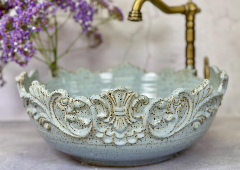 Puede incluir: Un lavabo de cer&aacute;mica azul claro con un borde adornado y decorativo. El lavabo tiene un esmalte moteado y un acabado ligeramente desgastado.