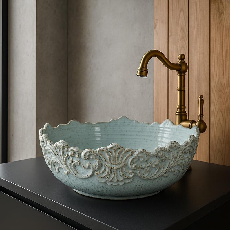 Puede incluir: Un lavabo de ba&ntilde;o de cer&aacute;mica azul claro con un dise&ntilde;o ornamentado y decorativo alrededor del borde. El lavabo est&aacute; sobre una encimera negra y un grifo dorado est&aacute; montado en la pared detr&aacute;s de &eacute;l.