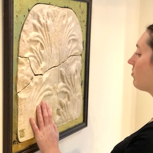May include: A white plaster relief sculpture of stylized plants framed in a dark brown frame with a gold-toned mat. The sculpture is mounted on a white wall. A person's hand is touching the sculpture.