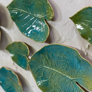 Puede incluir: Un conjunto de siete piezas de decoración de pared de hojas de cerámica en tonos verdes y azules. Las hojas son texturizadas y tienen un acabado brillante.