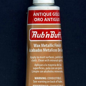 Peut inclure: Un tube de cire à finition métallique Rub'n Buff couleur or antique. Le tube possède un bouchon blanc et porte les inscriptions 'ANTIQUE GOLD ORO ANTIGUO', 'Wax Metallic Finish' et '1/2 fl oz (15 ml)'. L'emballage inclut des avertissements en anglais et espagnol.