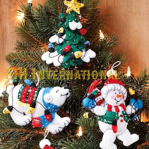 May include: Three felt Christmas ornaments featuring a green tree with a yellow star, a white polar bear with a red and blue blanket, and a white snowman wearing a red hat and green vest, all decorated with sequins and colourful light bulbs.