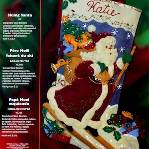May include: A Bucilla felt stocking kit titled Skiing Santa featuring a cream-colored stocking with a red-suited Santa Claus skiing, carrying a sack of toys, and decorated with sequins, beads, and the name Katie embroidered at the top.
