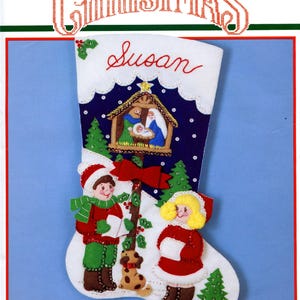 May include: A Bucilla Christmas felt stocking kit titled Away in a Manger, featuring a nativity scene, two children singing, and a small dog. The white stocking includes the name Susan in red script, green trees, and measures 18 inches diagonally.