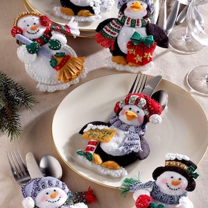May include: A set of six felt holiday cutlery holders featuring three penguins and three snowmen. The figures are decorated with scarves, hats, and festive items like a North Pole sign, a wreath, and a broom. Each holder is designed to tuck in silverware.