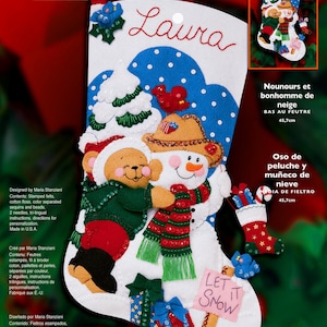 May include: A Bucilla felt Christmas stocking kit featuring a teddy bear and snowman design. The white stocking displays a bear hugging a snowman, decorated with sequins, beads, and a sign reading Let It Snow. The name Laura is embroidered at the top.