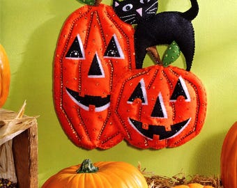 May include: A Bucilla felt craft kit for a wall hanging featuring two orange jack-o'-lanterns and a black cat. The finished size is 13.5 inches by 15.5 inches. The package includes stamped felts, cotton floss, sequins, beads, and needles.