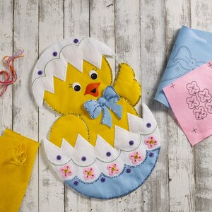 May include: A yellow felt chick hatching from a white and blue eggshell, decorated with purple beads, pink flower appliques, and a blue bow. The chick has black eyes and an orange beak, with additional felt fabric pieces nearby.