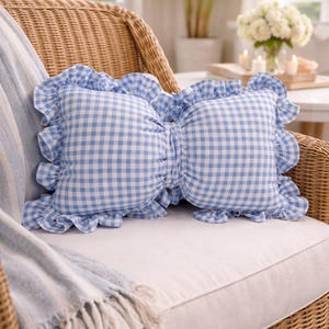 May include: Two blue and white gingham cushions with frilled edges sit on a white seat. The cushions are on a wicker chair, with a blue and white striped throw. The scene is bright and airy.