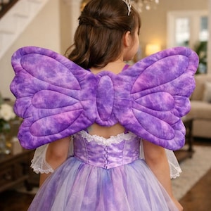 May include: A child wearing a purple fairy costume with large, padded butterfly wings. The dress is a gradient of purple and white, with a lace-trimmed bodice. A silver tiara is on the child's head.