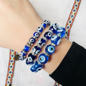 May include: Three blue beaded bracelets with evil eye designs. The bracelets feature round, blue beads with white and black eye details, interspersed with silver-coloured spacers. The bracelets are worn on a wrist, against a white shirt.