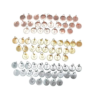 Puede incluir: Una colección de dijes metálicos circulares dispuestos en tres filas por color. La fila superior presenta dijes de oro rosa, la fila central dijes dorados y la fila inferior dijes plateados. Cada dije está grabado con una letra individual.