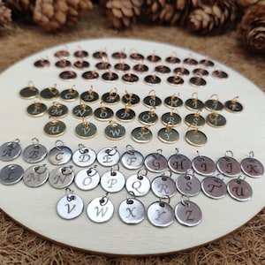 Peut inclure: Une collection de petits charms métalliques ronds, chacun gravé d'une lettre de l'alphabet. Les breloques sont en or, argent et cuivre, disposées sur une surface en bois. Le fond comprend des pommes de pin et une base en bois.