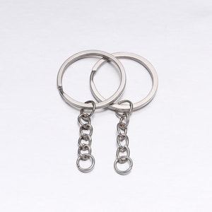 May include: Two silver-colored key rings with attached chains. Each ring is circular with a small opening. The chains are made of small, interconnected rings, ending in a smaller ring. The background is a light, metallic surface.