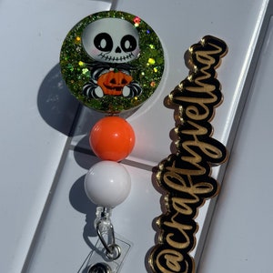 May include: A green and glitter Halloween themed badge reel with a skeleton holding a pumpkin. The reel has orange and white beads and a clear retractable badge holder. The text "@craftymama" is on a gold background.