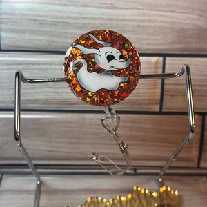 May include: A round, orange glitter badge holder with a white cartoon ghost with black eyes and a black mouth. The badge holder is attached to a clear plastic retractable badge reel.