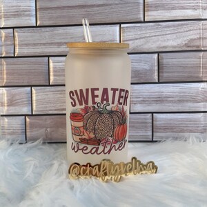 May include: A frosted glass tumbler with a bamboo lid and a clear straw. The tumbler features a fall-themed design with the words "Sweater Weather", a pumpkin, and a coffee cup. A gold-colored sign with the text "@craftyvelma" is in front of the tumbler.