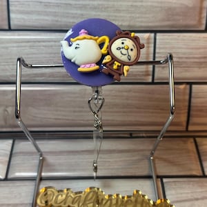 May include: A purple and white round badge with a cartoon image of Mrs. Potts and Cogsworth from Beauty and the Beast. The badge is attached to a silver retractable badge reel with a clear plastic strap. The badge reel is hanging from a gold metal stand with the word "craftyvetra" written on it.
