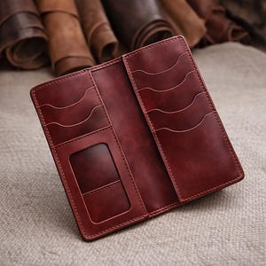 May include: Open burgundy leather wallet with several card slots and a clear ID window. The rectangular wallet has a stitched border. Rolls of leather are visible in the background.