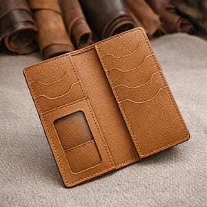 May include: Open, tan leather wallet with multiple card slots and a clear ID window. The wallet is stitched with a lighter thread and has a textured surface. Rolls of leather are visible in the background.