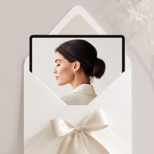 May include: A white envelope with a cream-colored bow holds a tablet displaying a woman with dark hair and gold earrings. The envelope is mounted on a light gray background, with delicate white floral accents.