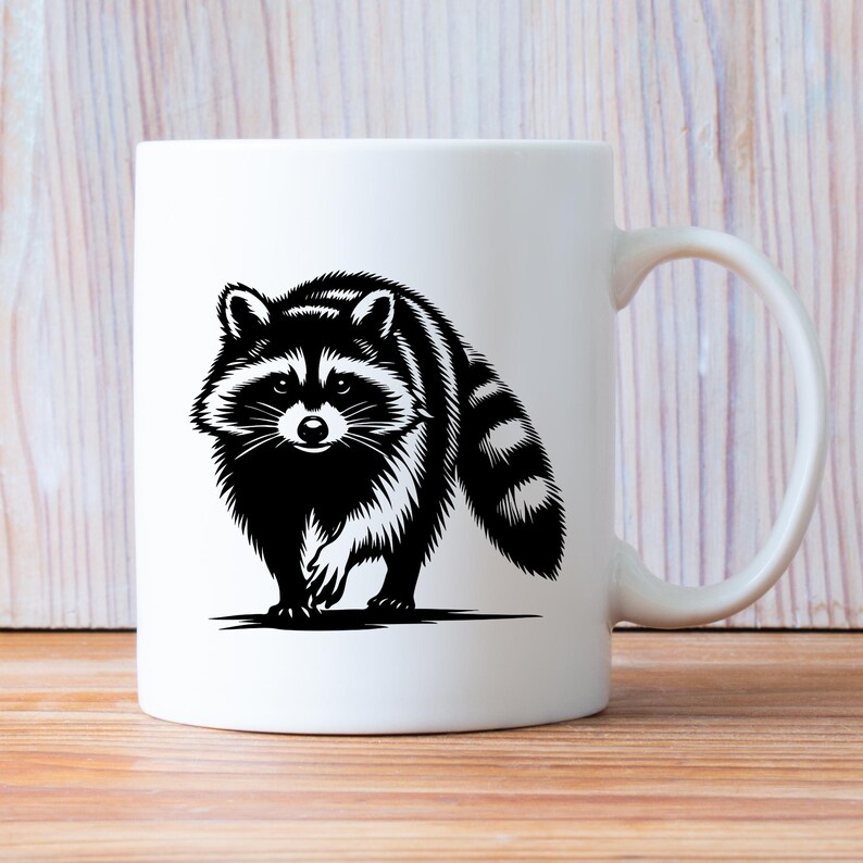 May include: White ceramic coffee mug with a black and white illustration of a raccoon. The raccoon is facing forward with its head tilted slightly to the side. The raccoon has a bushy tail and a black mask around its eyes.