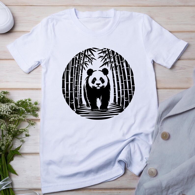 May include: White t-shirt featuring a black and white panda bear design. The panda is standing in a bamboo forest, enclosed in a circular frame. The shirt is made of a soft material and has short sleeves.