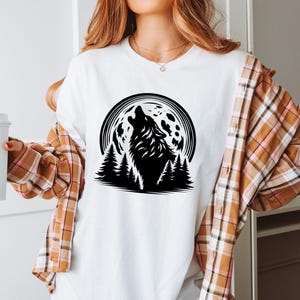May include: A white t-shirt with a black silhouette of a wolf howling at the moon. The moon is full and has a textured surface. The wolf is surrounded by a forest of pine trees.