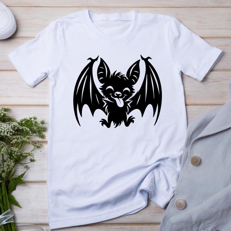K&ouml;nnte beinhalten: Wei&szlig;es T-Shirt mit einem schwarzen Cartoon-Fledermaus-Design. Die Fledermaus hat die Zunge herausgestreckt und die Fl&uuml;gel ausgebreitet. Das T-Shirt ist aus weichem Material. Geeignet f&uuml;r Halloween.