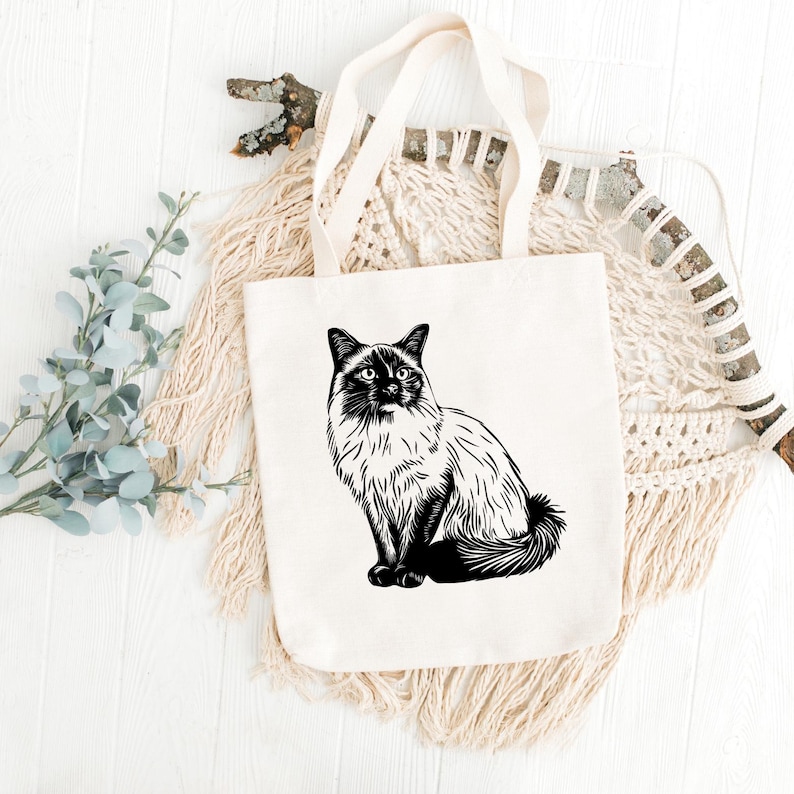May include: A white tote bag featuring a black and white illustration of a Siamese cat sitting on the front. The cat has blue eyes and a long tail.