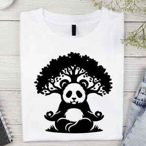 May include: White t-shirt featuring a black graphic of a panda meditating under a tree. The panda is in a lotus position, with the tree's branches forming a canopy above its head. The design is centered on the shirt.