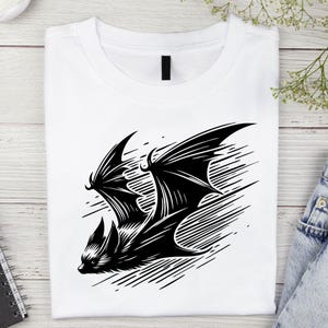 K&ouml;nnte beinhalten: Wei&szlig;es T-Shirt mit einer schwarzen Fledermaus-Illustration. Die Fledermaus ist im Flug mit detaillierten Fl&uuml;geln und K&ouml;rper dargestellt. Das Design befindet sich vor einem Hintergrund aus diagonalen Linien. Das T-Shirt ist sauber gefaltet.