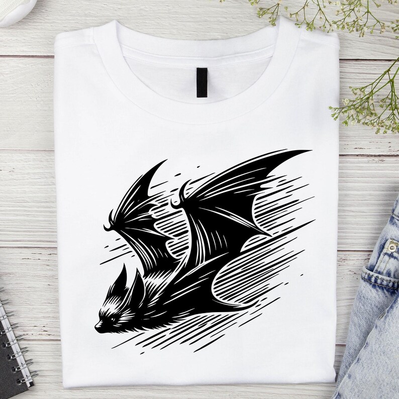 K&ouml;nnte beinhalten: Wei&szlig;es T-Shirt mit einer schwarzen Fledermaus-Illustration. Die Fledermaus ist im Flug mit detaillierten Fl&uuml;geln und K&ouml;rper dargestellt. Das Design befindet sich vor einem Hintergrund aus diagonalen Linien. Das T-Shirt ist sauber gefaltet.