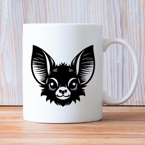 K&ouml;nnte beinhalten: Wei&szlig;e Keramiktasse mit einer schwarz-wei&szlig;en Illustration eines Chihuahua-Gesichts. Das Design zeigt die gro&szlig;en Ohren, die runden Augen und den kleinen, l&auml;chelnden Mund des Hundes. Die Tasse hat einen Standardgriff und eine glatte, gl&auml;nzende Oberfl&auml;che.