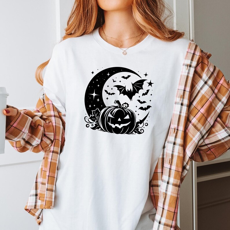 K&ouml;nnte beinhalten: Wei&szlig;es T-Shirt mit einem schwarzen Halloween-Design. Die Grafik zeigt einen Halbmond, Flederm&auml;use, Sterne und einen geschnitzten K&uuml;rbis. Teil einer Halloween-Kollektion.