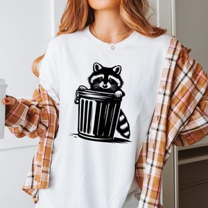 May include: A white t-shirt with a black and white illustration of a raccoon peeking out of a trash can.