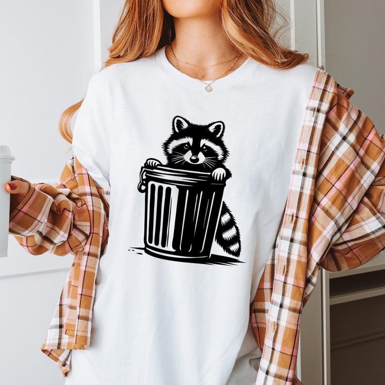 May include: A white t-shirt with a black and white illustration of a raccoon peeking out of a trash can.
