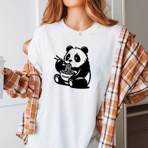 May include: White t-shirt featuring a black and white panda bear graphic. The panda is depicted eating noodles from a bowl with chopsticks. The shirt has short sleeves and a crew neck.