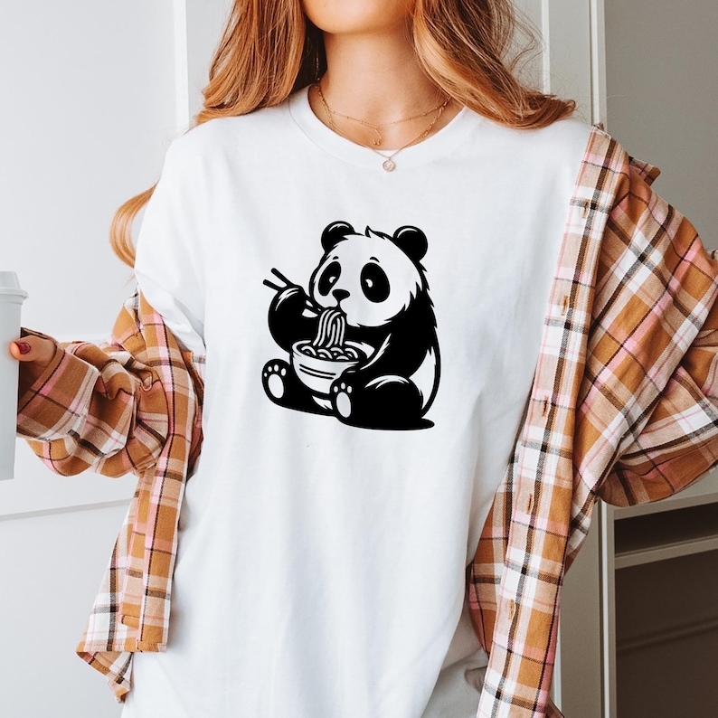 May include: White t-shirt featuring a black and white panda bear graphic. The panda is depicted eating noodles from a bowl with chopsticks. The shirt has short sleeves and a crew neck.