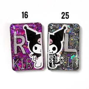 Puede incluir: Dos dijes rectangulares de resina etiquetados 16 y 25. El dije izquierdo es morado con purpurina, presenta un personaje Kuromi y las letras R y KIR. El derecho es negro con purpurina, presenta un Kuromi esqueleto y las letras L y KIR.
