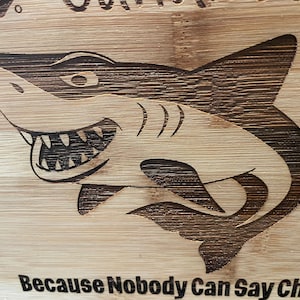 Puede incluir: Una tabla de cortar de madera con un gr&aacute;fico de tibur&oacute;n y el texto "Because Nobody Can Say Char-c..."