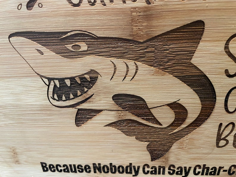 Puede incluir: Una tabla de cortar de madera con un gr&aacute;fico de tibur&oacute;n y el texto "Because Nobody Can Say Char-c..."