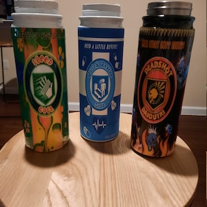 May include: Three colorful insulated beverage containers with different designs. The first container is green, yellow, and orange with the text "Speed Up Your Life" and "Cola". The second container is blue and white with the text "Need a Little Revive!" and "Quick Revive Soda". The third container is black with orange and yellow flames and the text "Deadshot Daiquiri".