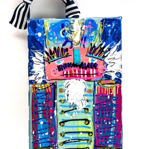May include: A colorful, abstract painting of a nutcracker soldier. The artwork features a pink face, blue and purple accents, and a teal jacket with gold buttons. A black and white striped ribbon is attached at the top.