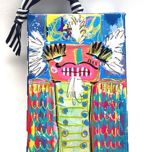 May include: A colorful, abstract painting of a nutcracker soldier. The artwork features a pink face, a green jacket, and a blue background. The painting is adorned with white accents and a black and white striped ribbon for hanging.