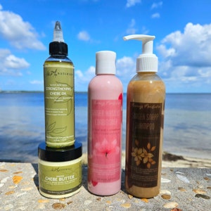 May include: Three hair care products on a stone surface with a blue sky and water in the background. The products are a brown bottle labeled "Scalp and Hair Strengthening Chebe Oil", a pink bottle labeled "Ginger Hibiscus Conditioner", and a brown bottle labeled "Ginger Snapp Shampoo".